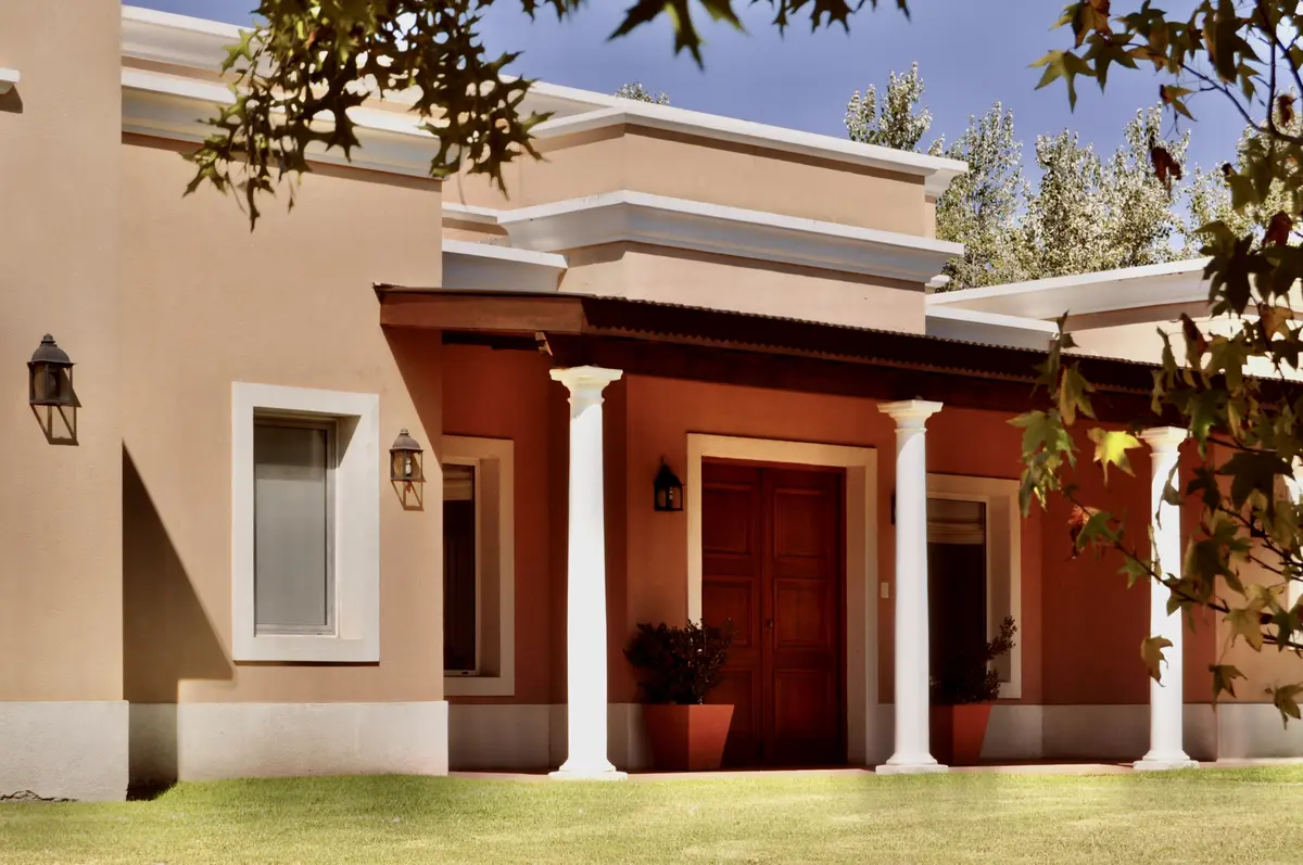 Fachada contemporánea con frente rojo, columnas blancas y jardín verde — proyecto Chacras de la Cruz, por Marconi Silva Arquitectos, en Capilla del Señor