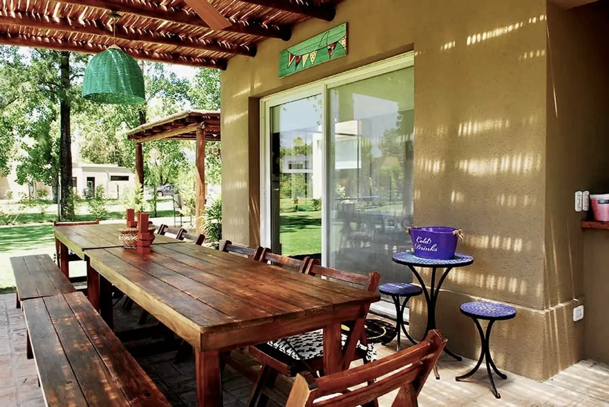 Galería rústica con mesa madera, pérgola y vista al jardín — proyecto El Encuentro  2, por Marconi Silva Arquitectos, en Benavidez