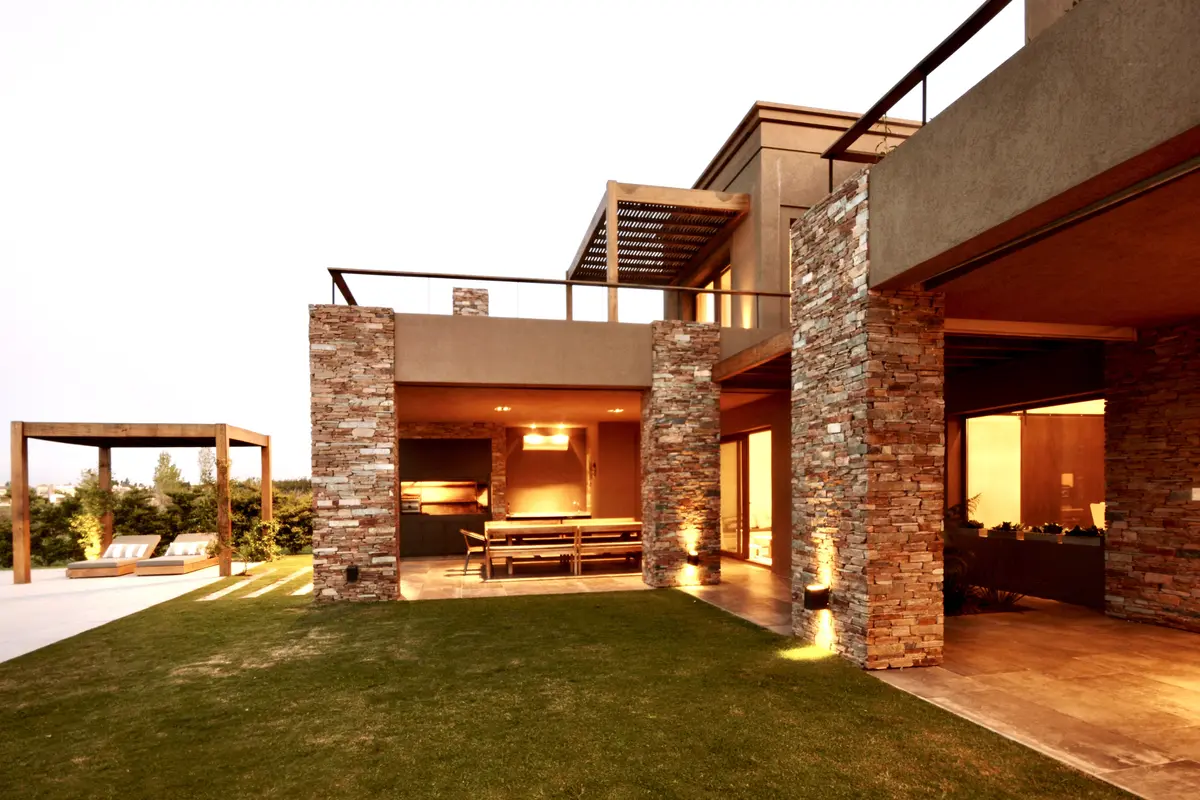 Casa contemporánea con patio, césped y pilares de piedra iluminada — proyecto Golf Club Nordelta, por Marconi Silva Arquitectos, en Nordelta