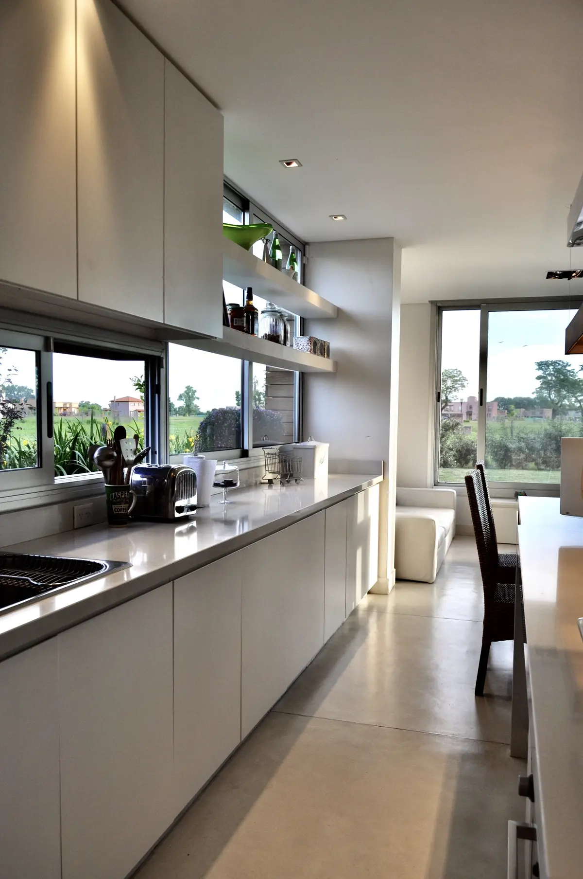 Cocina minimalista blanca con vista al campo, mesada de mármol gris y ventanales — proyecto Haras Santa María, por Marconi Silva Arquitectos, en Escobar