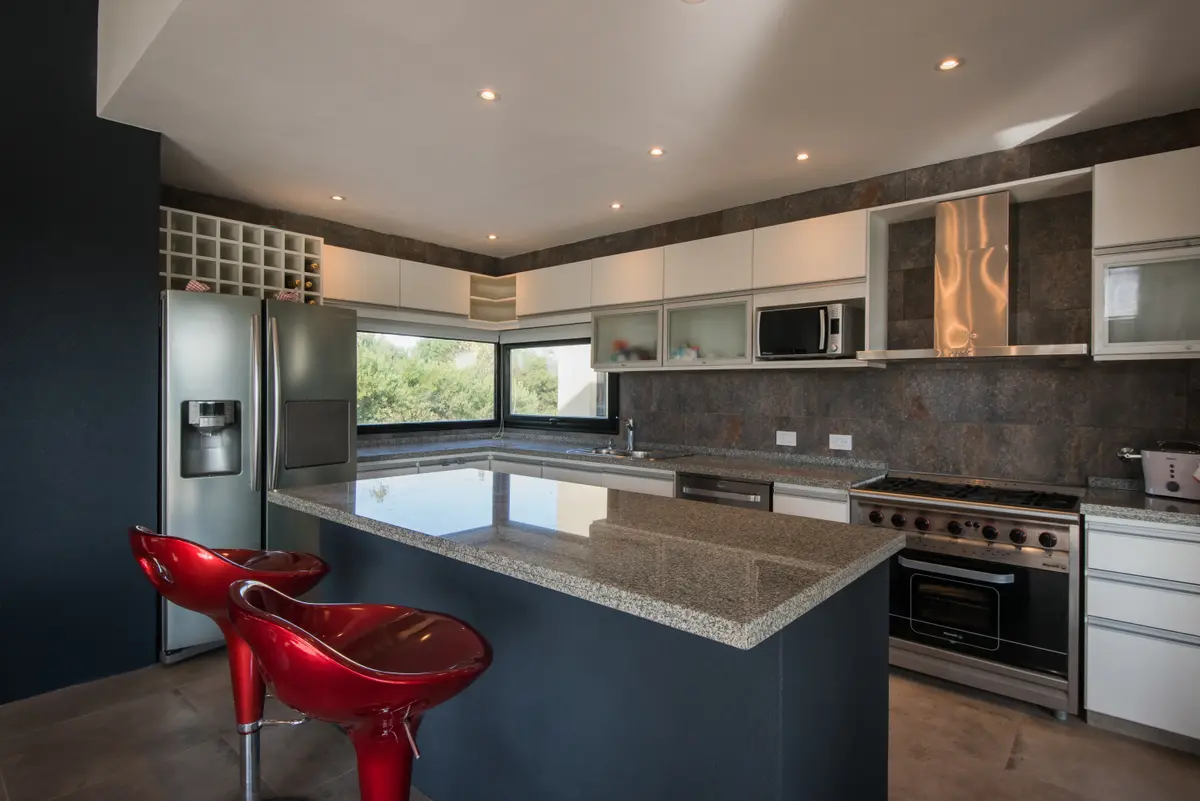 Cocina moderna con isla grande, mesada de piedra y banquetas rojas — proyecto Costa Esmeralda 3, por Arquitecto Caceres, en Costa Esmeralda