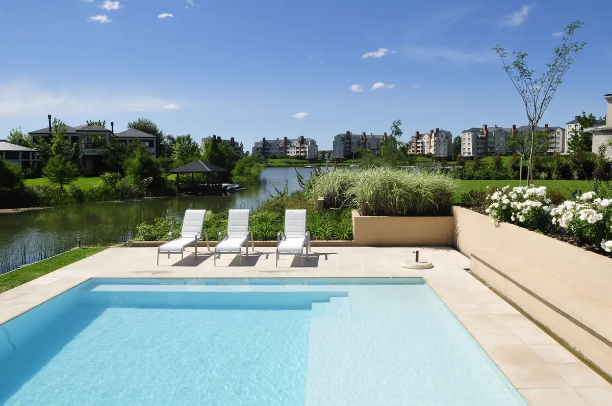 Piscina moderna con reposeras blancas junto a un lago en un día soleado — proyecto Al agua, por Ricardo Pereyra Iraola , en Nordelta