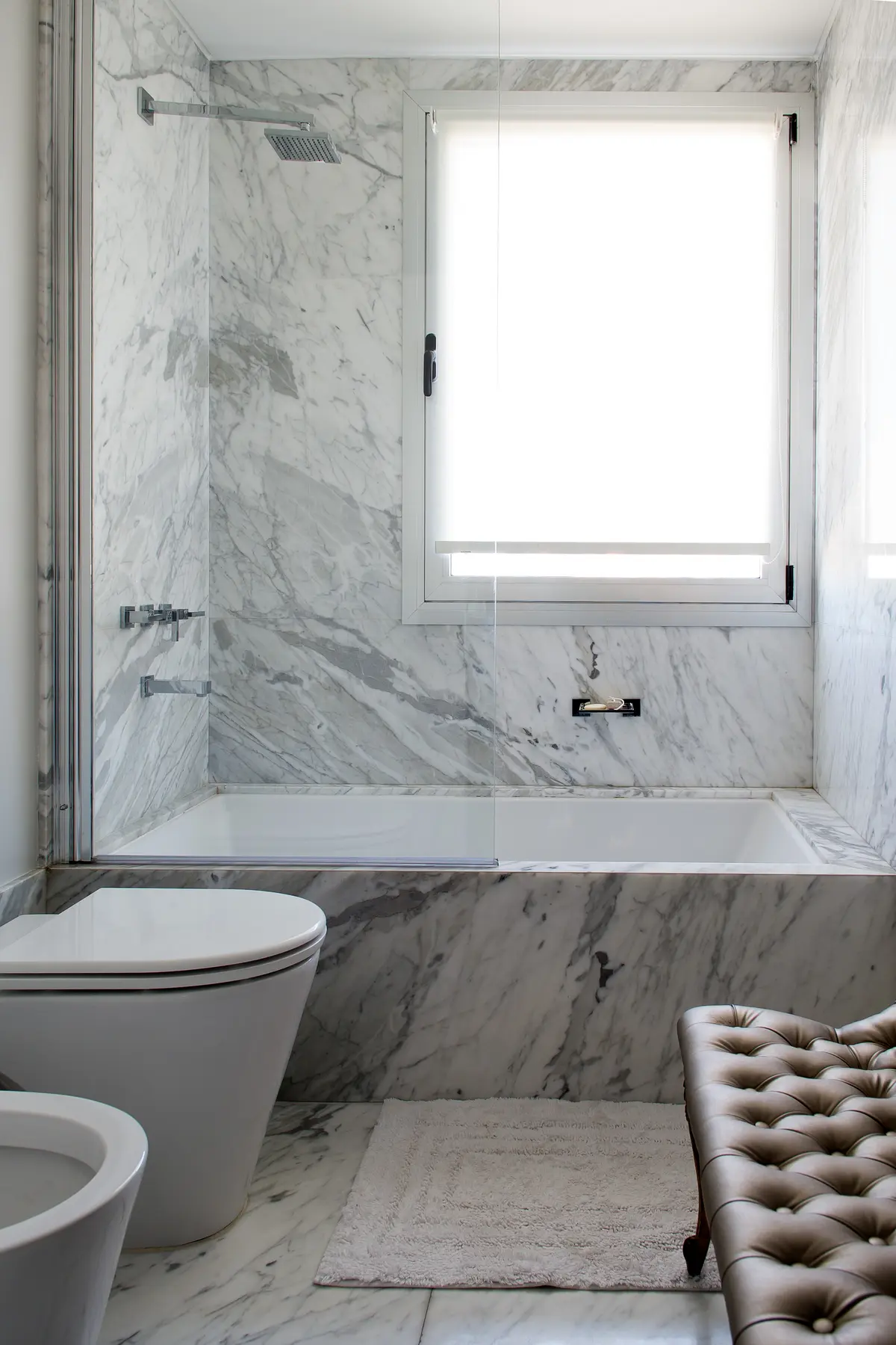 Baño moderno revestido en mármol con bañera, inodoro y banqueta capitoné — proyecto Libertador y Lafinur, por Estudio Magrane, en CABA