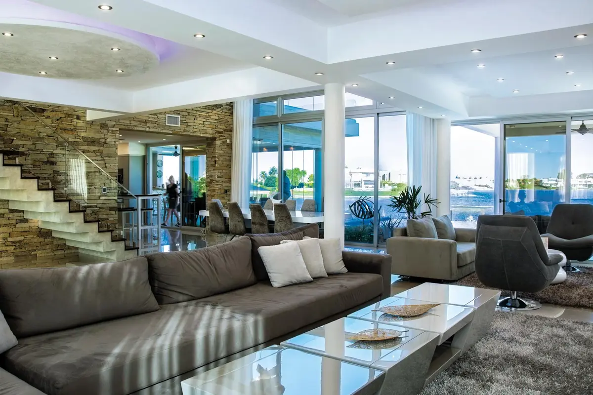 Living moderno con ventanales y detalles en piedra y madera — proyecto La Reserva Cardales I, por ARQCONS, en Los Cardales