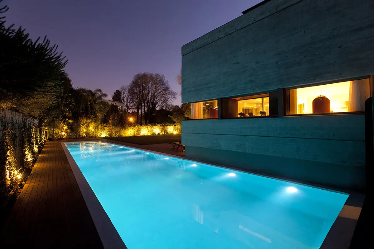 Piscina iluminada moderna junto a fachada de hormigón — proyecto Acasusso, por Remy Arquitectos, en Acassuso