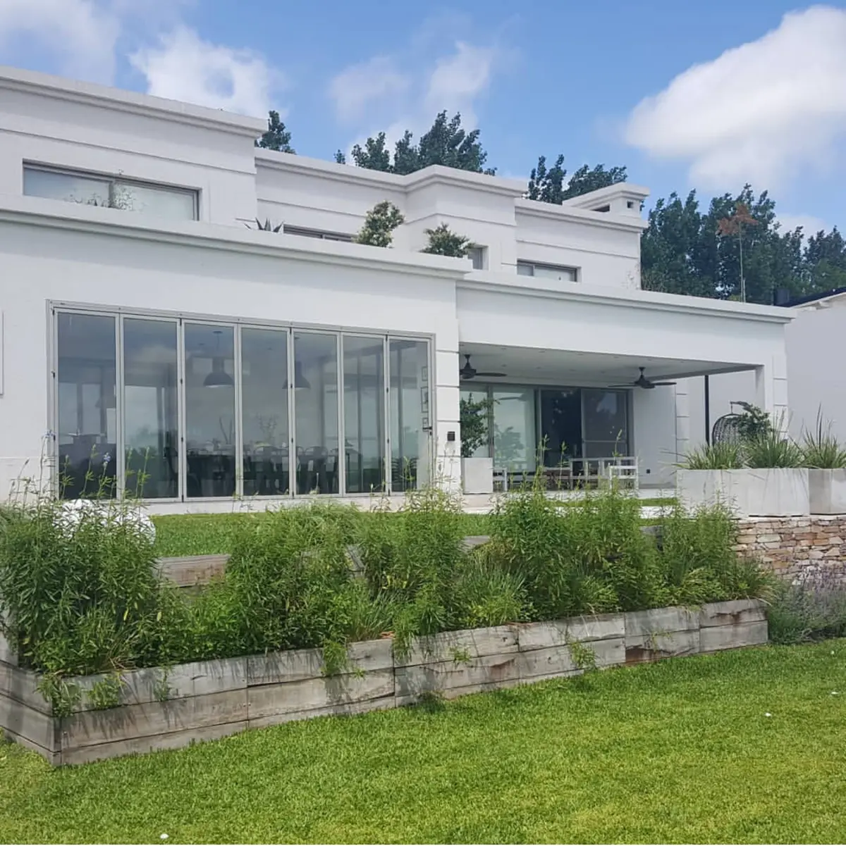 fachada moderna blanca con grandes ventanales y jardin exuberante — proyecto LRCC 249, por LESSI ARQUITECTURA, en Los Cardales