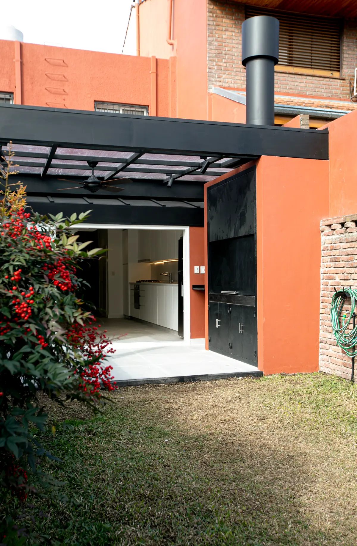 Área de churrasco moderna com fachada laranja, pergolado preto e vegetação exuberante — projeto Obra Martinez A, por Arq. Lourdes Yarade Saravia, em Martínez
