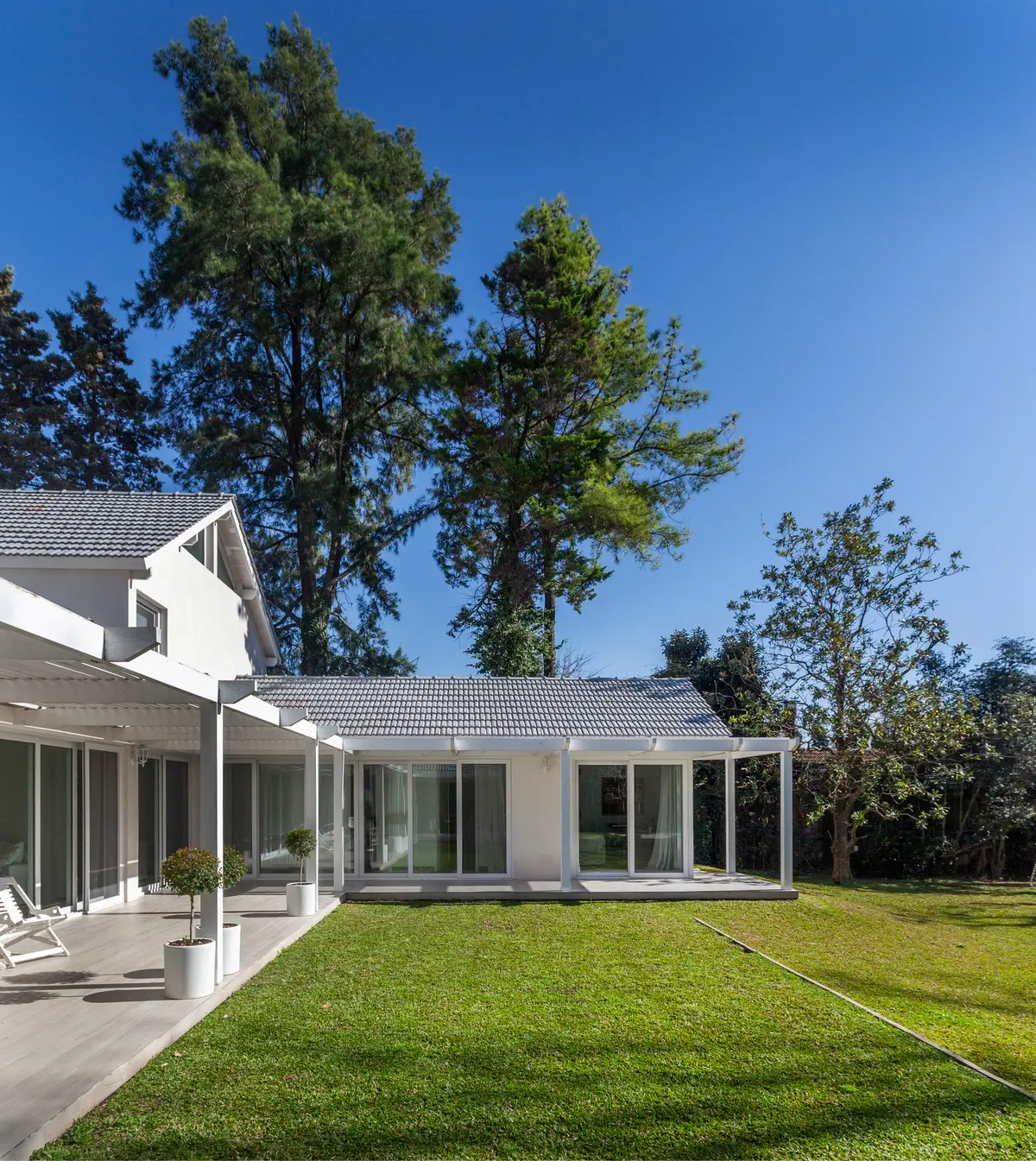 Casa moderna blanca con césped amplio y árboles altos — proyecto Highland, por Lala Loviscek arquitecta, en Pilar