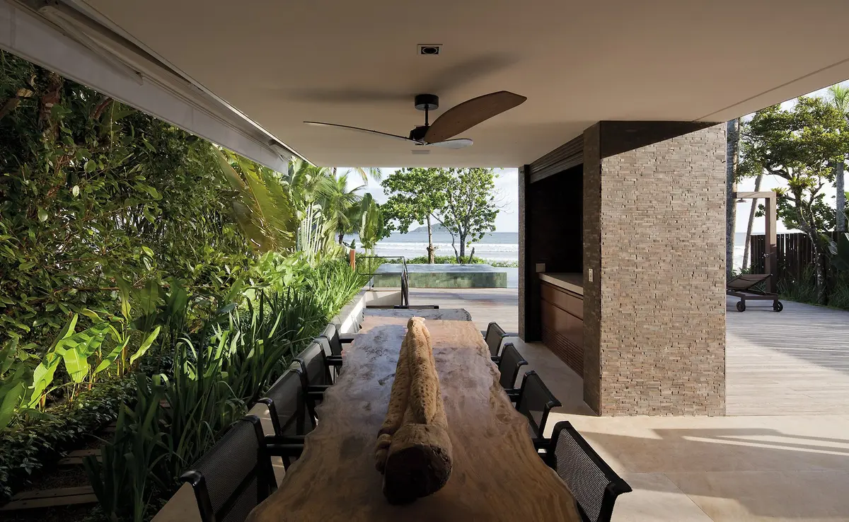 Galería cubierta con jardín vertical, mesa de madera y vista a pileta — proyecto West Whales, por Gil Fialho