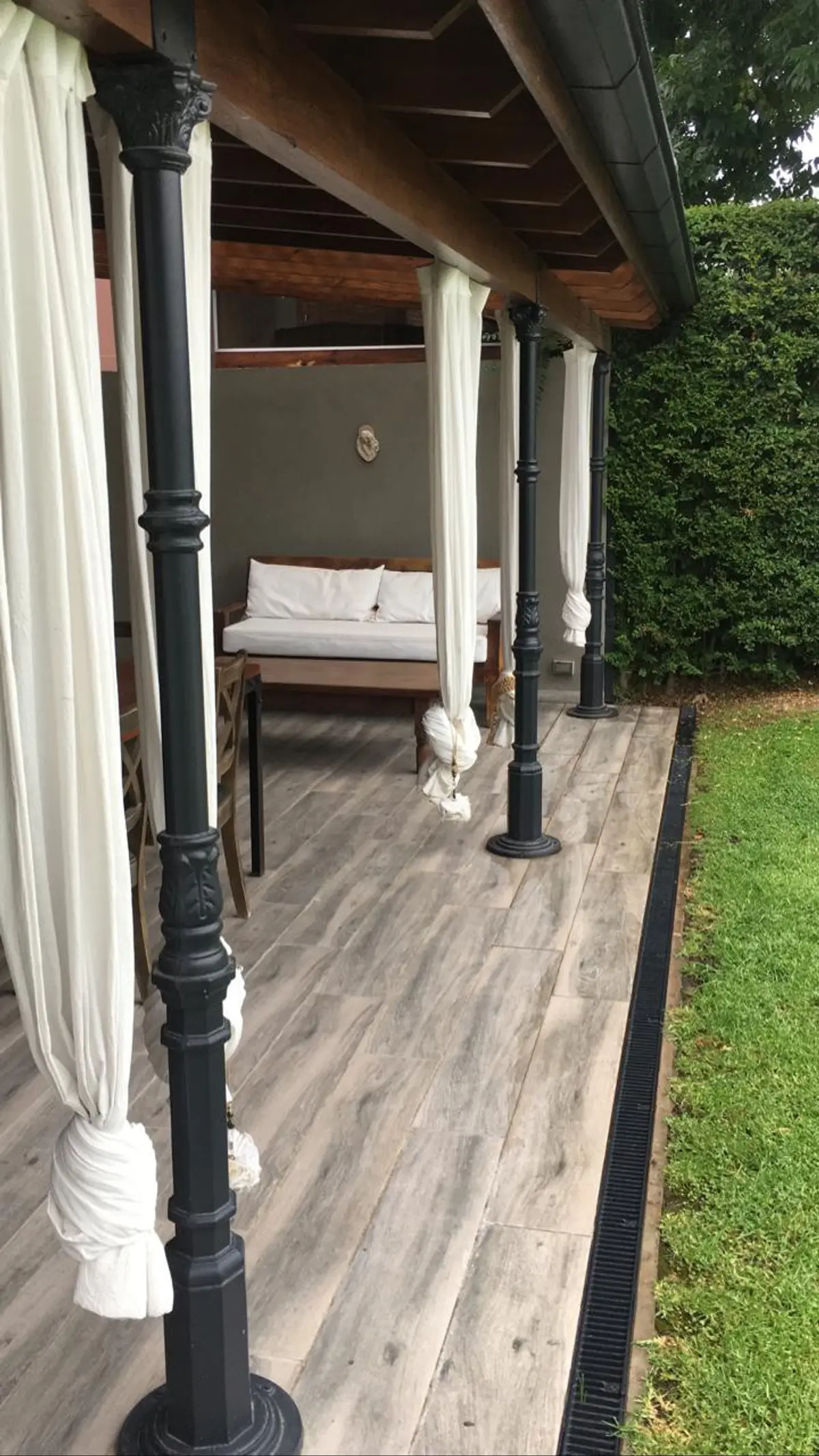 Galería techada con columnas negras, cortinas blancas y cama de madera — proyecto Pilar, por Rioja Arquitectura, en Pilar