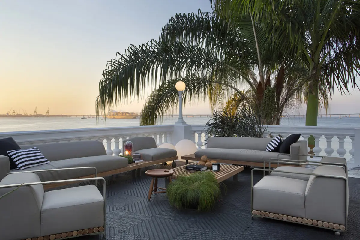 Terraza de lujo con sofás grises, frente a bahía y palmeras al atardecer — proyecto Casa COR 2019, por Studio Ro-ca