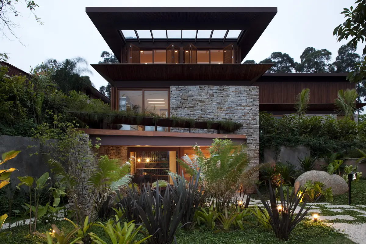 Casa contemporánea con fachada de piedra, grandes ventanales y jardín frondoso iluminado — proyecto Residência MN, por Gil Fialho
