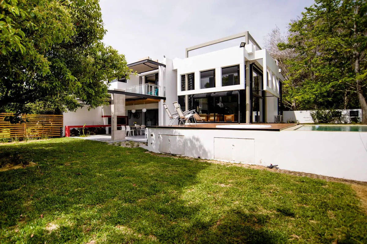Fachada moderna blanca con grandes ventanales y jardín exuberante — proyecto CASA GONNET, por LEONE LORAY ARQUITECTOS, en Gonnet