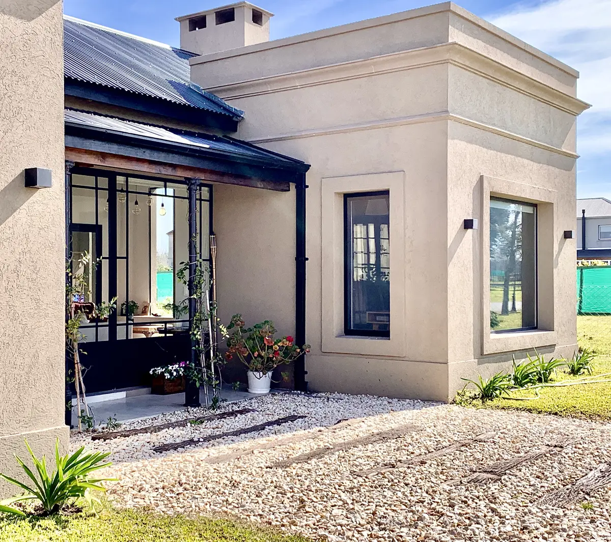 Fachada moderna color beige con grandes ventanales negros y jardín con grava — proyecto Los Jazmines Pilar del Este, por Marconi Silva Arquitectos, en Pilar del Este