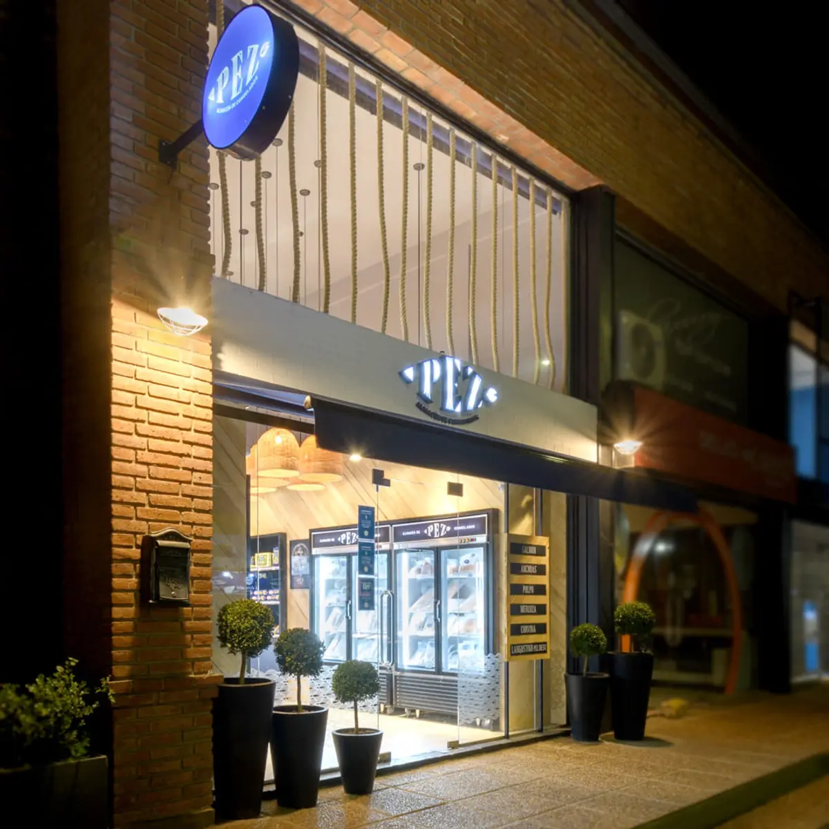 Fachada nocturna de local comercial con ladrillo y vidrieras iluminadas — proyecto PEZ, Almacén de Congelados - Arquitectura Comercial, por Síncresis Arquitectos, en Ciudad de Córd