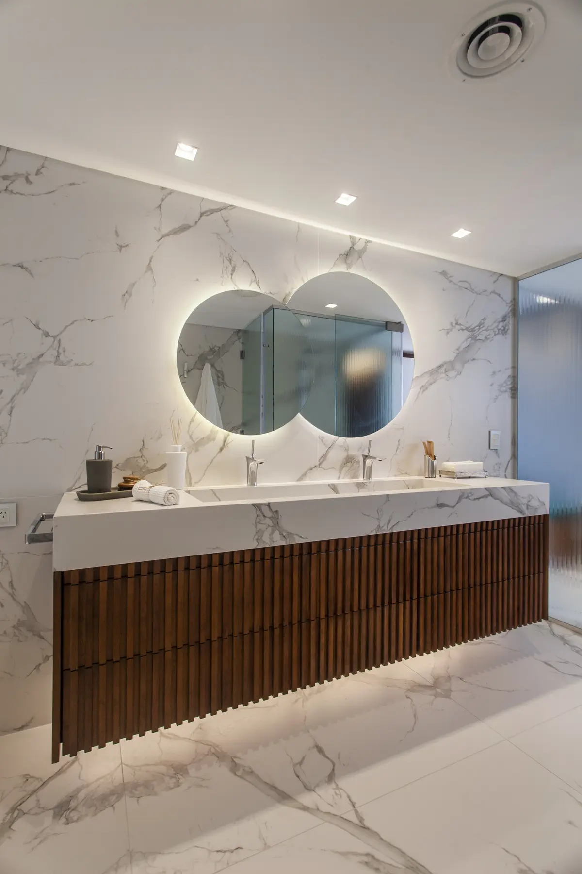 Baño moderno con doble lavabo, espejo curvo retroiluminado y mueble de madera — proyecto Pacífico BRT, por Vanguarda Arquitectos, en CABA