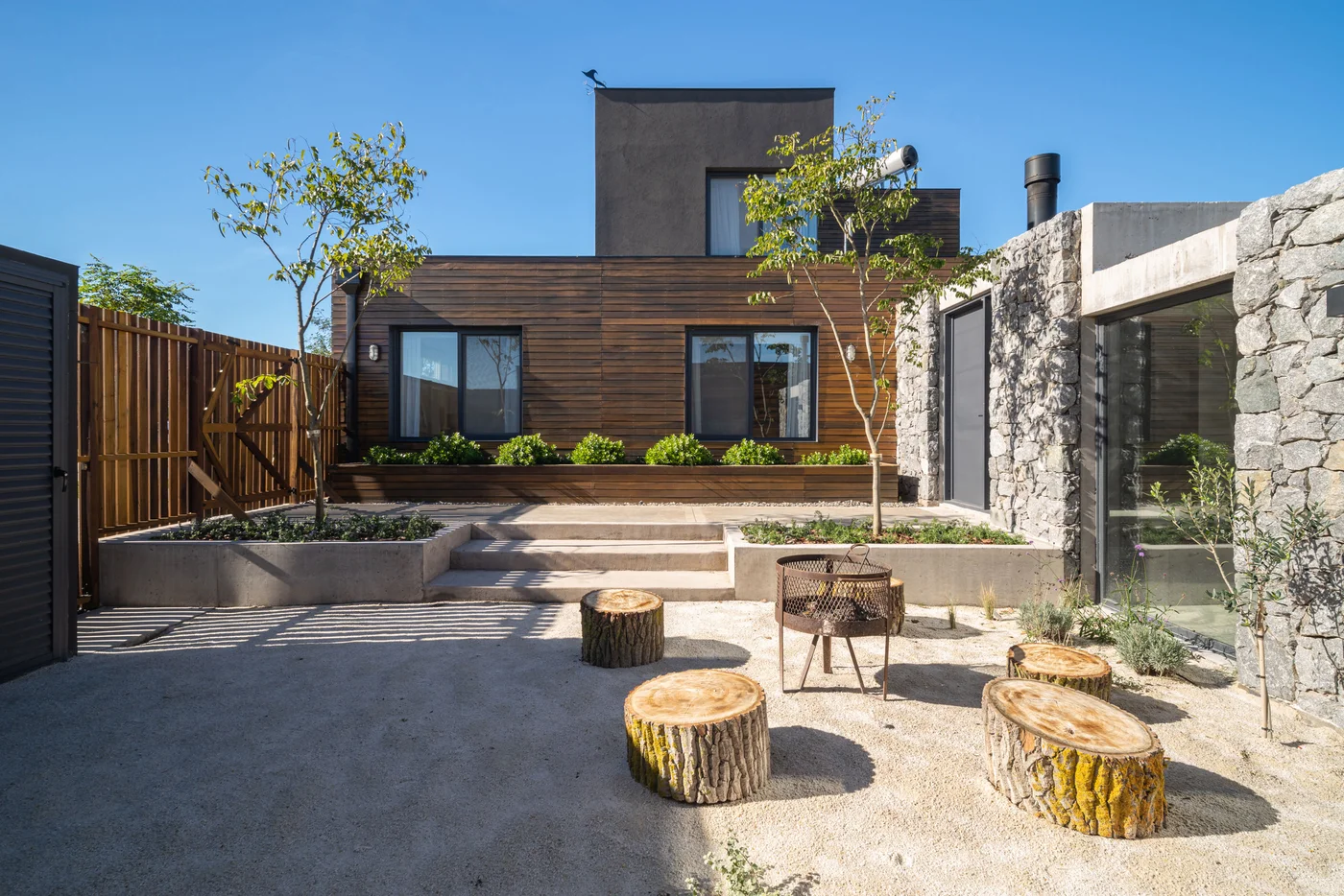 Fachada moderna con piedra y madera, patio con troncos como asientos.