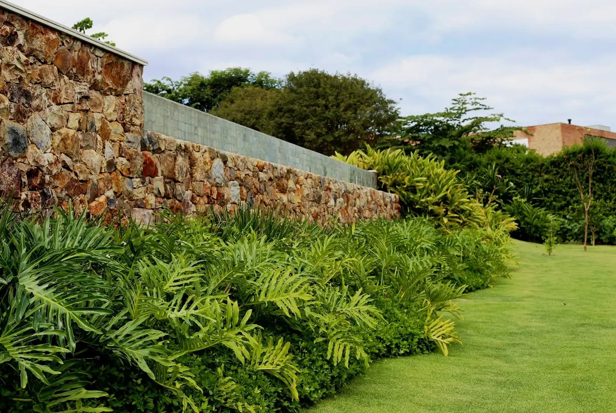 Jardín moderno con muro de piedra, plantas y césped verde impecable — proyecto Residência CA, por Gil Fialho
