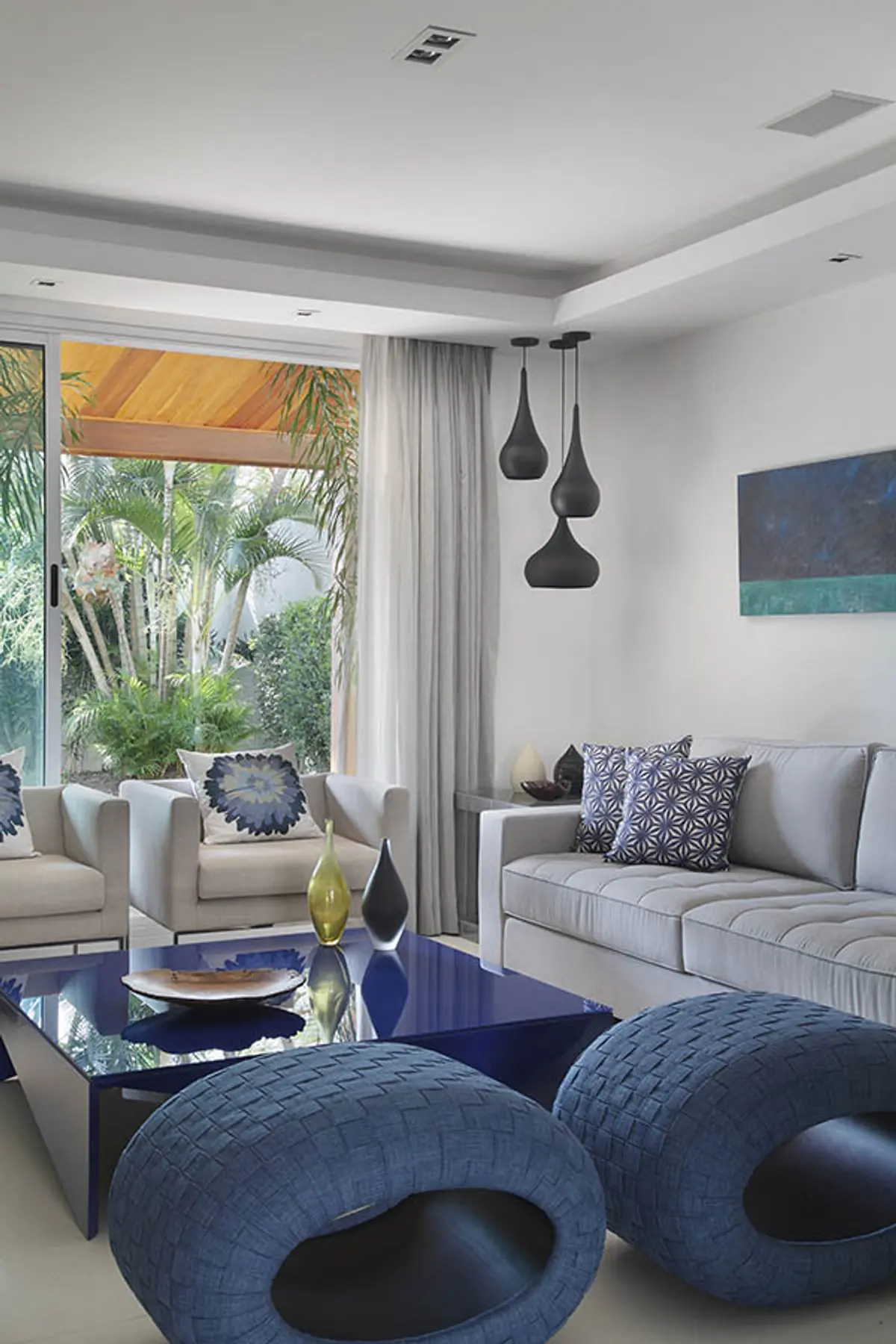 Living contemporáneo con puffs azules, sofá gris y vista al jardín tropical — proyecto Casa Barra ED, por Amanda Miranda