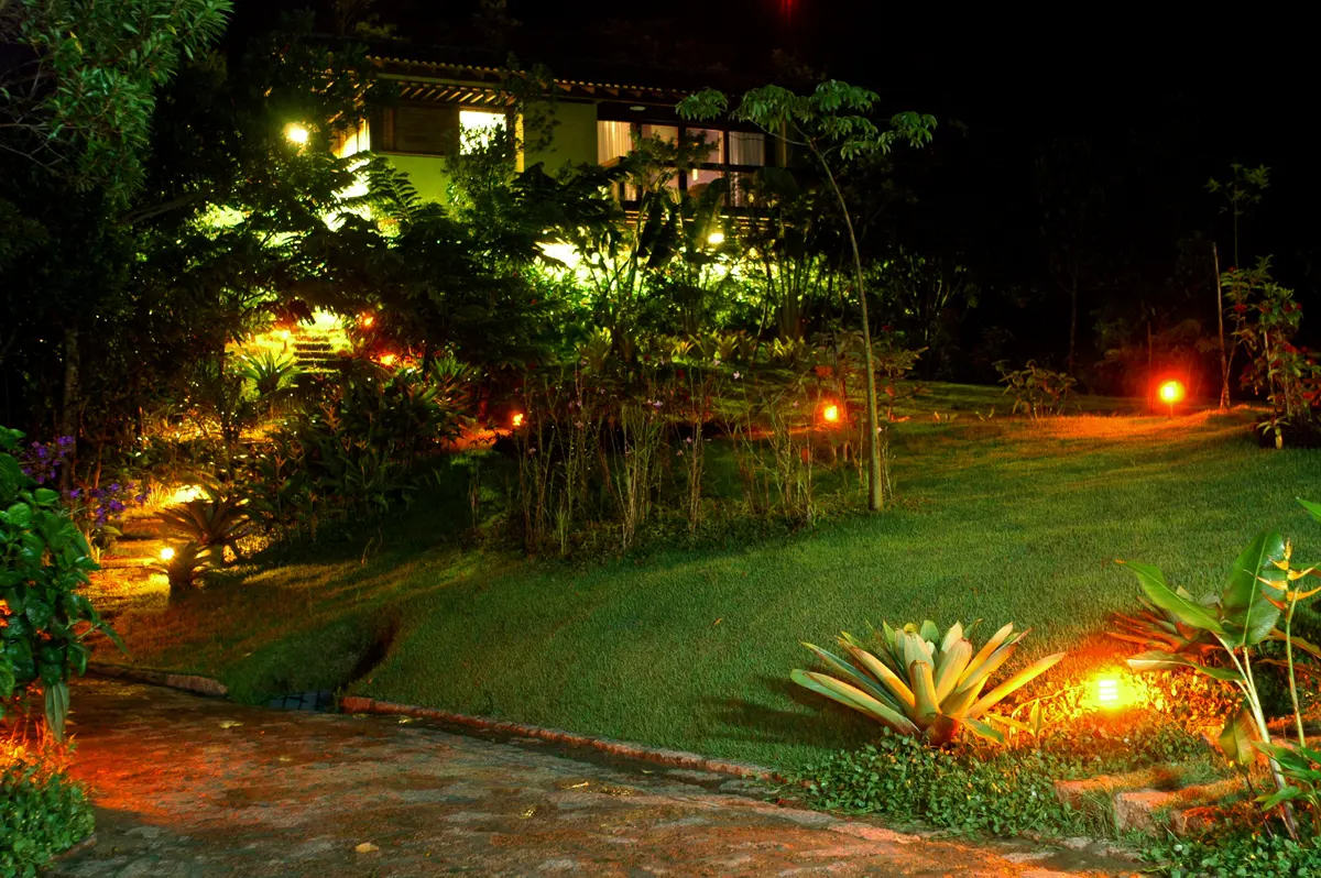 Jardín nocturno iluminado con casa, árboles y deck de madera — proyecto Fazenda Indaiauba, por Gil Fialho