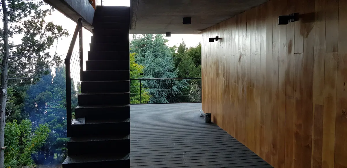 Escada flutuante de concreto preto, deck cinza e parede de madeira em exterior florestal — projeto Casa Península San Pedro, por Servente Larrazabal Arq, em Bariloche