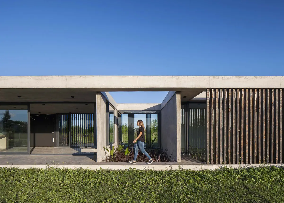 Casa moderna com fachada de concreto e madeira, pátio e vegetação — projeto Chacra 93, por V2 Arquitectos, em City Bell