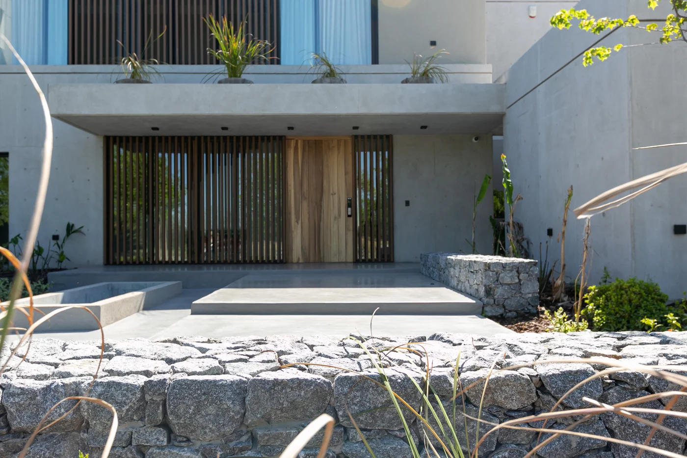 Fachada moderna de hormigón y madera con puerta pivotante y jardineras de piedra.