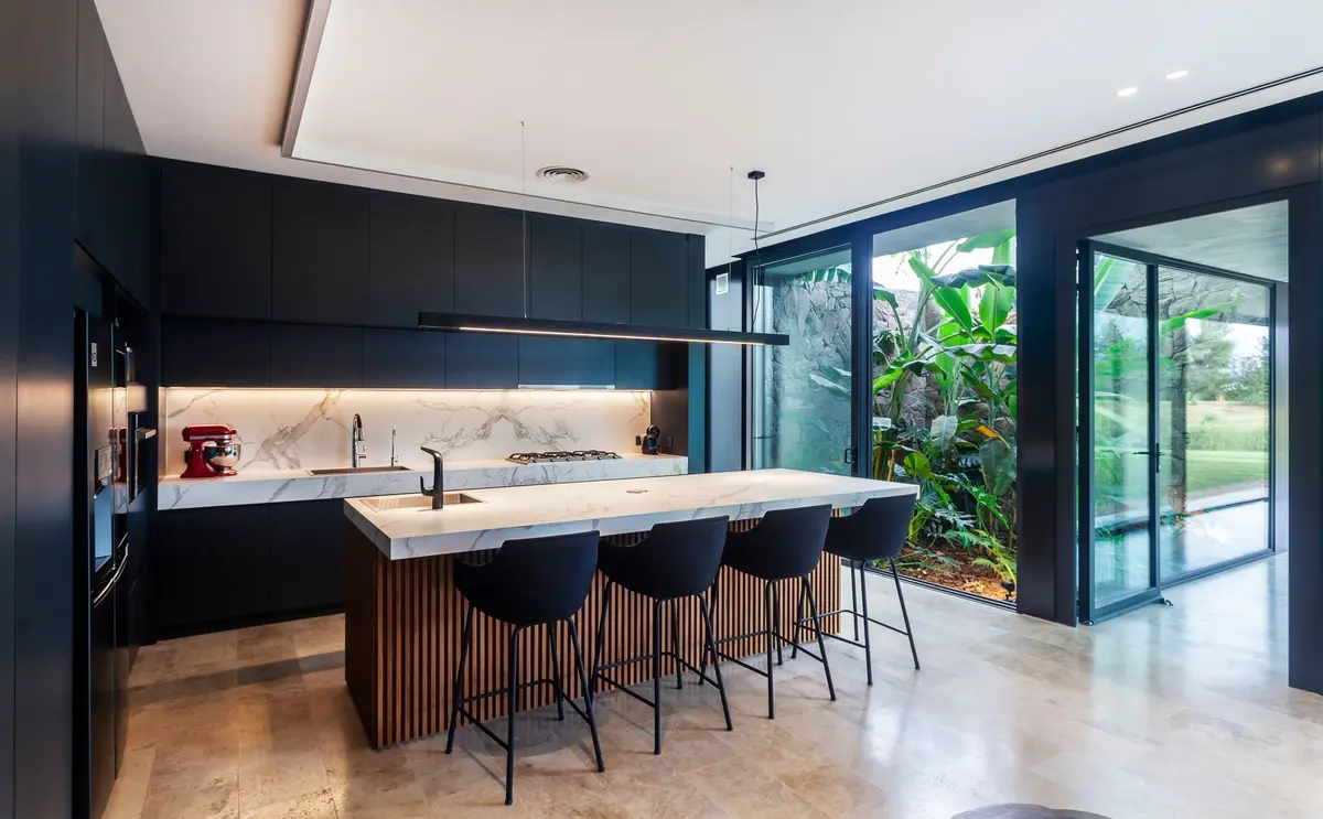 Cozinha moderna com ilha grande, janelas do chão ao teto e vegetação exuberante — projeto BUNKER, por OON ARCHITECTURE