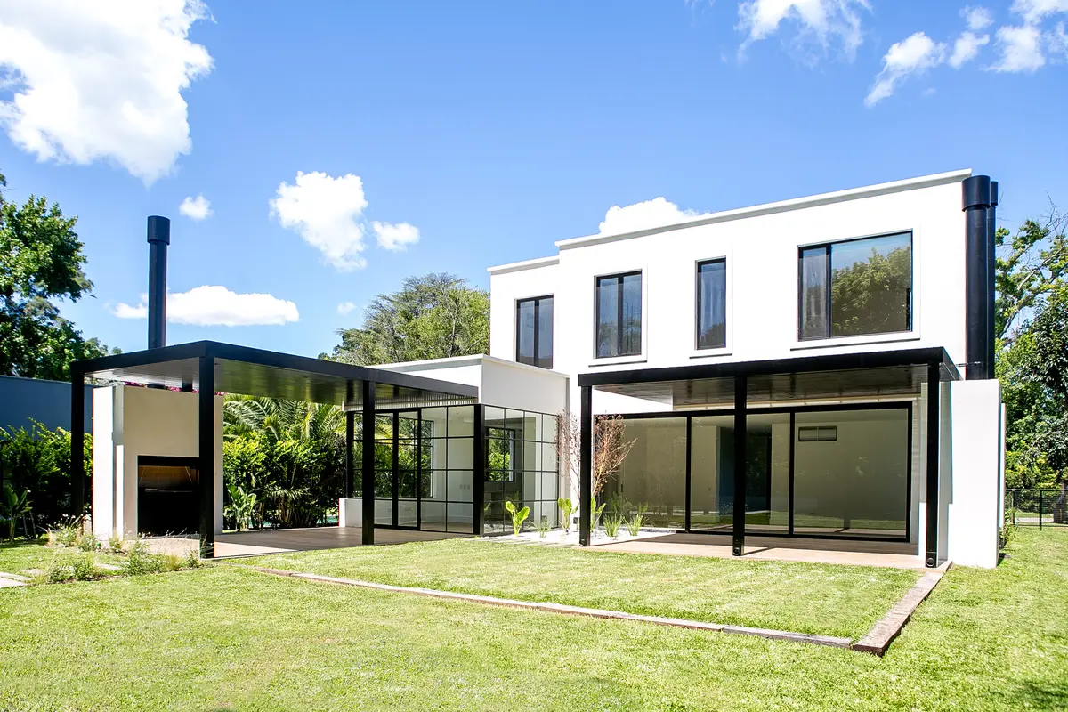 Casa moderna blanca con pérgola negra, jardín y parrilla exterior — proyecto Bosque Chico, por Estudio Arq. María Sioli, en Garin
