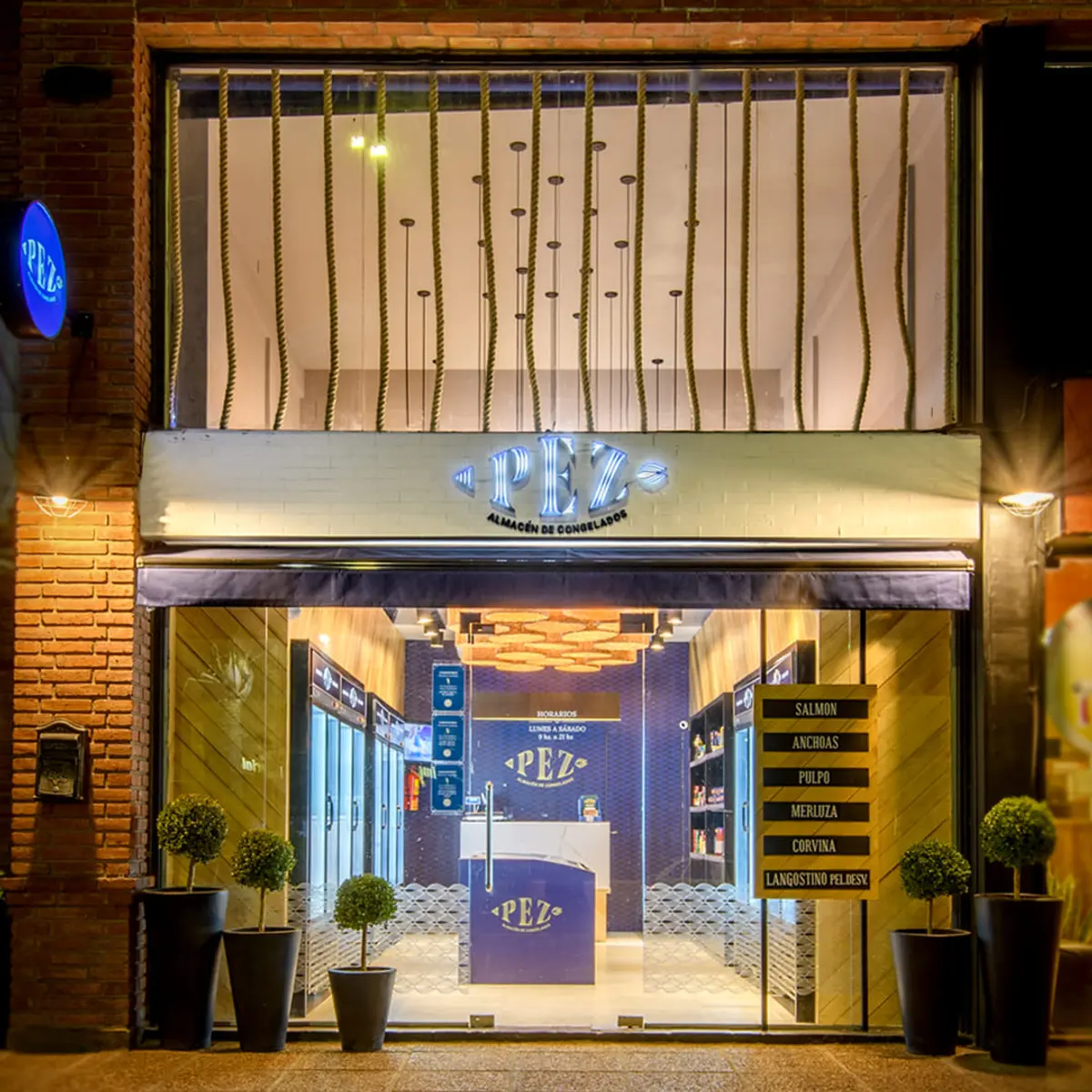 Fachada nocturna de local comercial con vidrieras y plantas decorativas — proyecto PEZ, Almacén de Congelados - Arquitectura Comercial, por Síncresis Arquitectos, en Ciudad de Córd