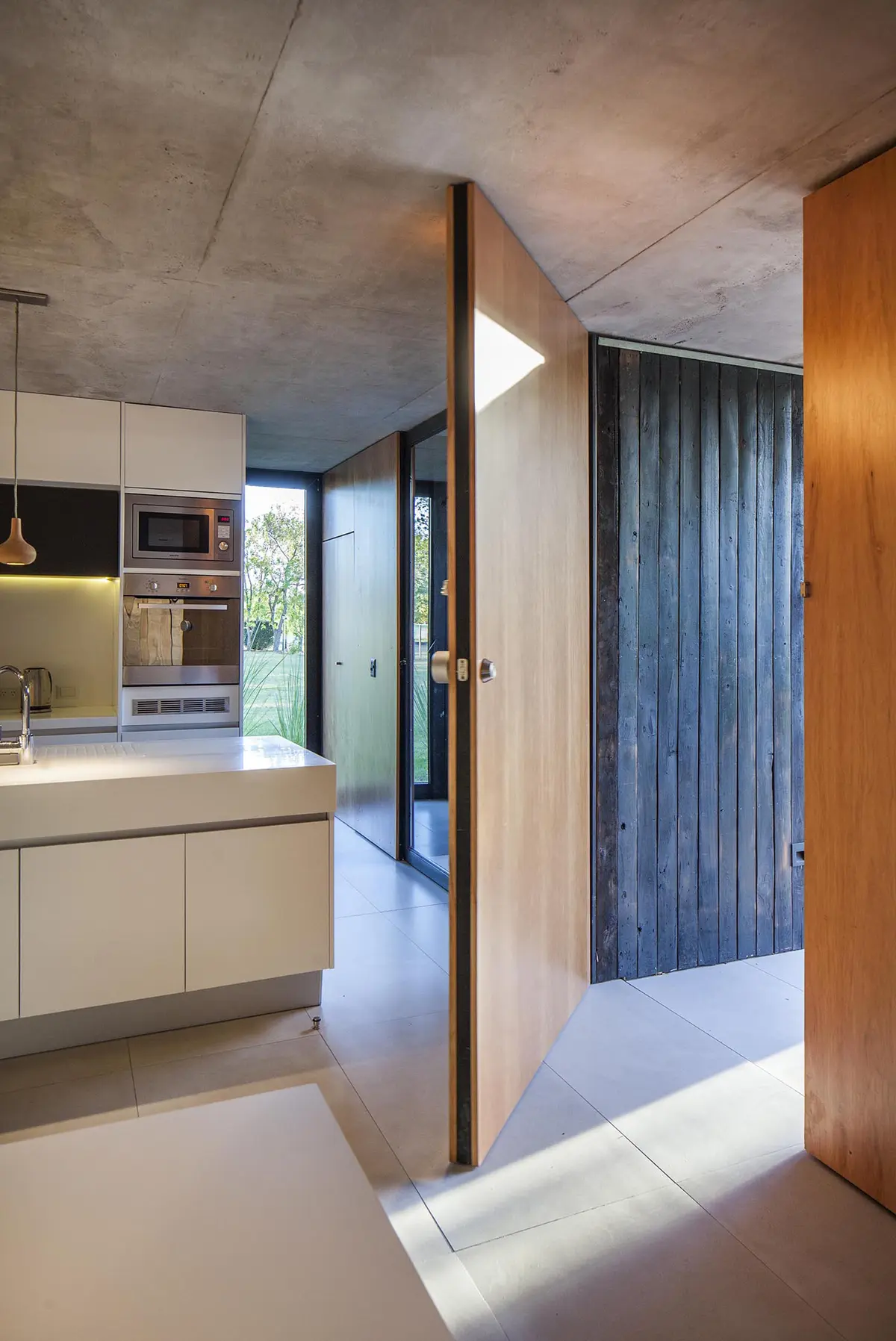 Cocina moderna con puerta pivotante y techo de hormigón expuesto — proyecto Casa MF, por V2 Arquitectos, en City Bell