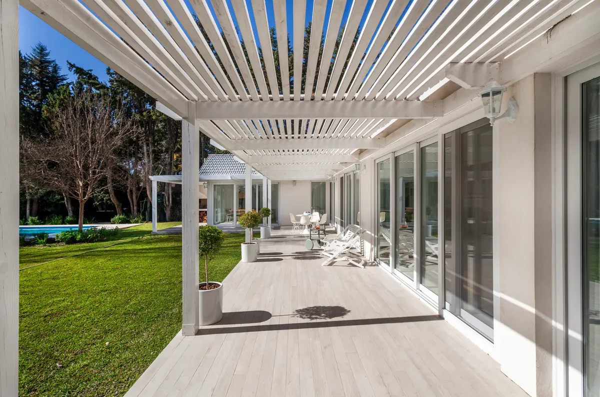 Pérgola blanca con terraza de madera y césped frondoso — proyecto Highland, por Lala Loviscek arquitecta, en Pilar