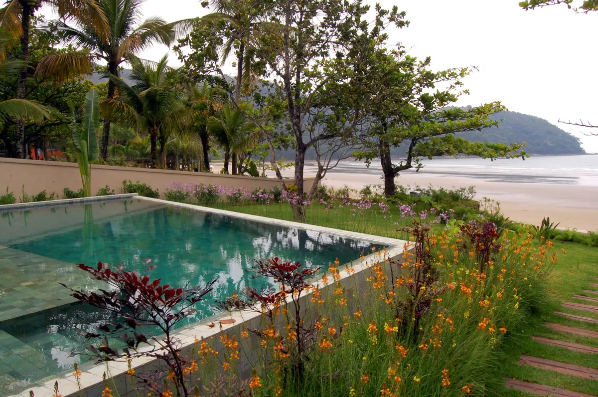 Pileta verde turquesa frente a playa con palmeras y jardín — proyecto West Whales, por Gil Fialho